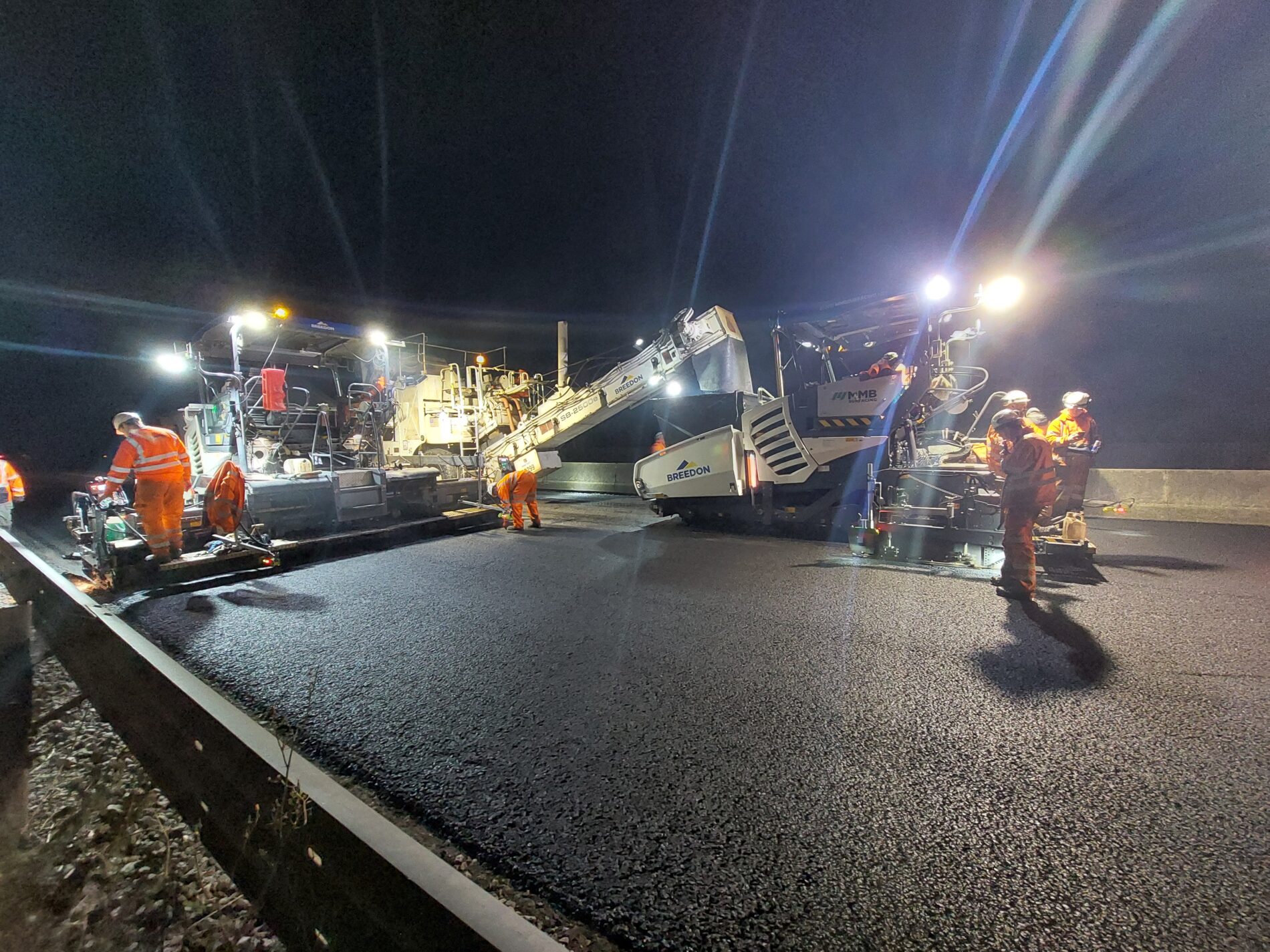 Night-time road resurfacing works on a motorway, with a Breedon paving machine laying fresh asphalt while workers in high-visibility orange suits oversee the operation